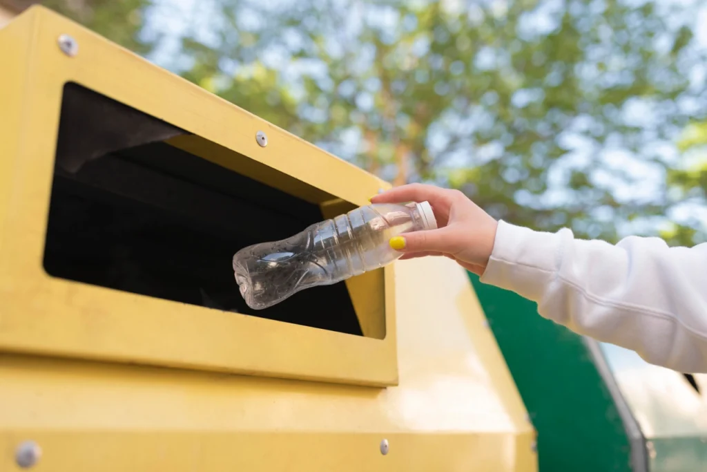 kobieta wrzuca plastikową butelkę do żółtego kontenera w punkcie selektywnej zbiórki odpadów komunalnych